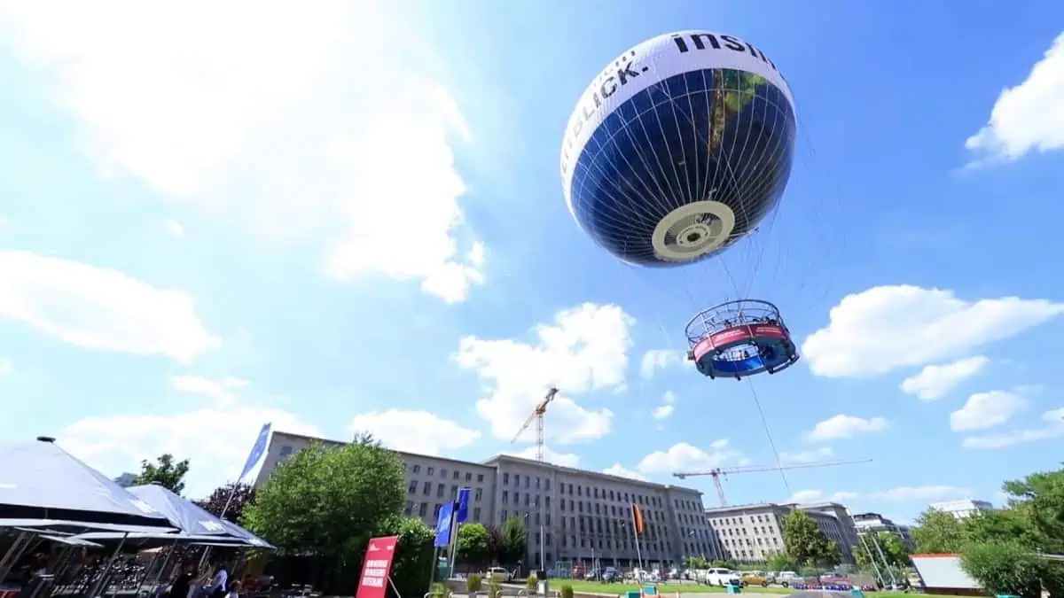 World-Balloon-with-Perfect-View Tethered World Balloon in Berlin rising above city buildings with passengers in the circular viewing platform against a bright blue sky